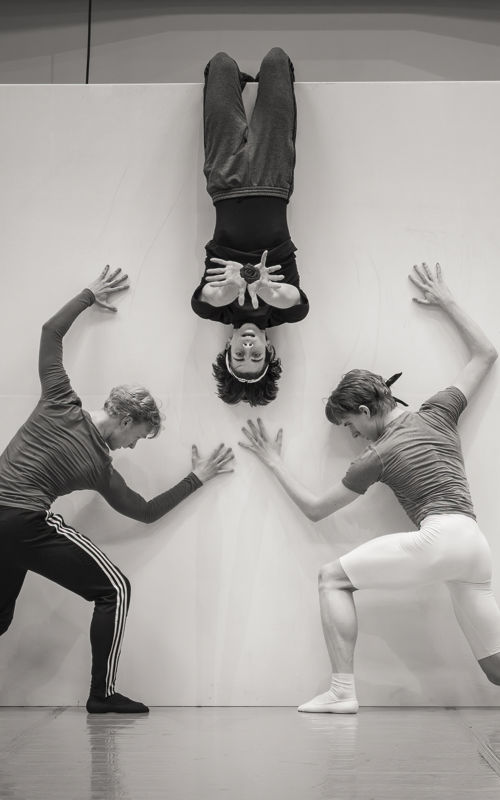 en danser henger opp ned på en vegg - og to andre holder hendene mot den samme hvite veggen