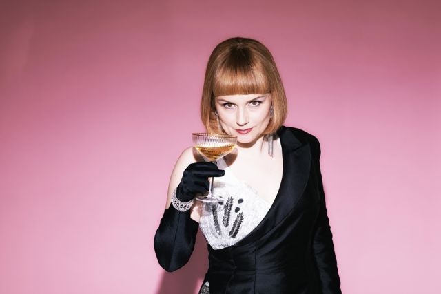 Violetta i La traviata ser ned med et glass champagne i hånden. Kjolen og hår og sminke er i art -deco-stil