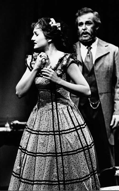 Gammelt svart-hvitt bilde fra operaforestilling, kvinne i kjole og mann i bakgrunnen