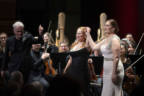 Dirigent og to kvinner i konsertkjoler smiler mot publikum