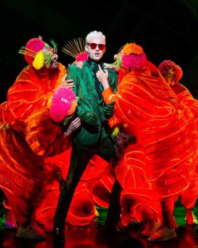 En mann i grønt kostyme er omkranset av skikkelser i neon-røde kostymer