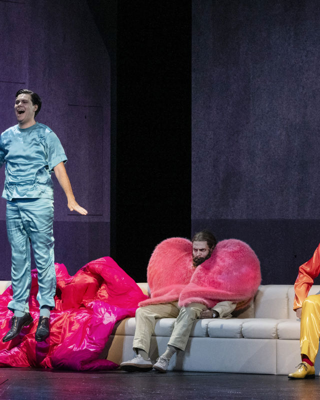 Mann i blå trøye og bukse hopper foran sofa, der det sitter en mann med hjertejakke og en med gul-oransje dress