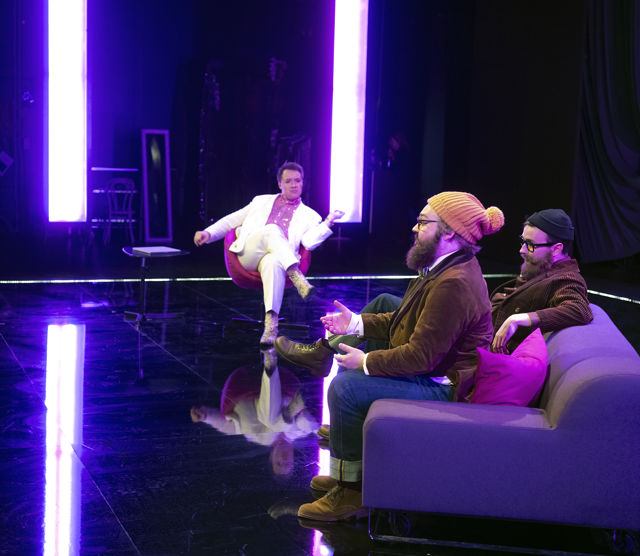 Mann i hvit dress intervjuer to menn i sofa