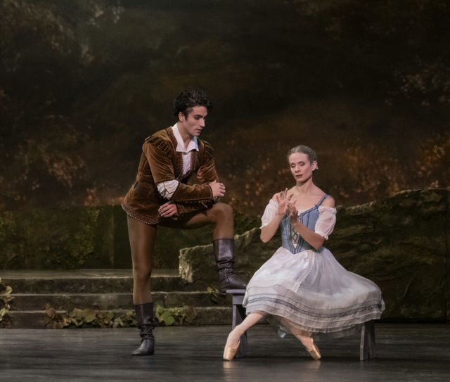 To ballettdansere på scene med skogsbakgrunn; én står med benet på en trapp i brun kostyme, den andre sitter i hvit og blå drakt.