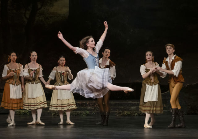 Ballettdanser i lyseblå og hvit drakt hopper i grand jeté foran seks dansere i jordfargede kostymer på scene med skogsbakgrunn.