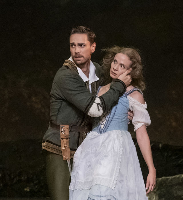 To ballettdansere står på en mørk scene. Den ene danseren holder den andre nært, og begge har kostymer fra balletten Giselle.