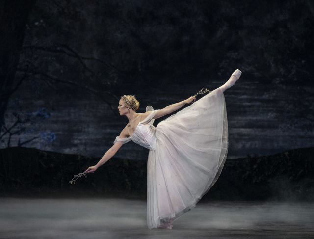 Ballettdanser i hvit tutu står i arabesque med blomster i hånden på mørk, tåkete scene.
