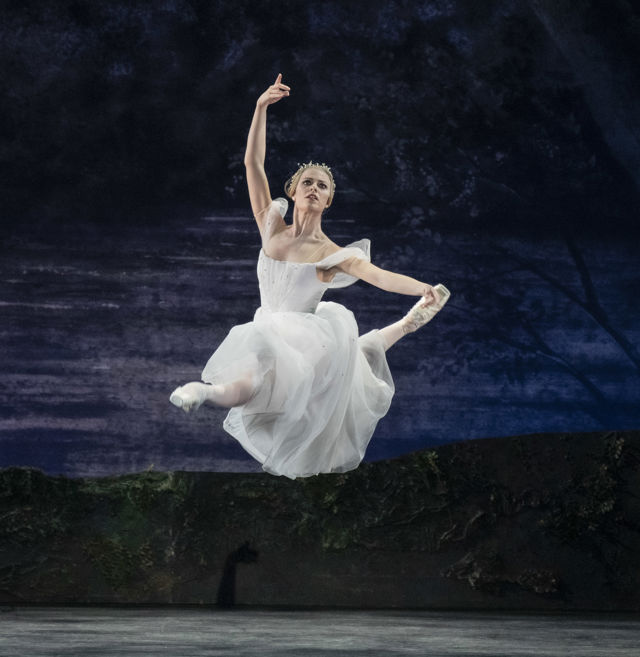 Ballettdanser i hvit tutu hopper med armene hevet på mørk scene med skogsbakgrunn.