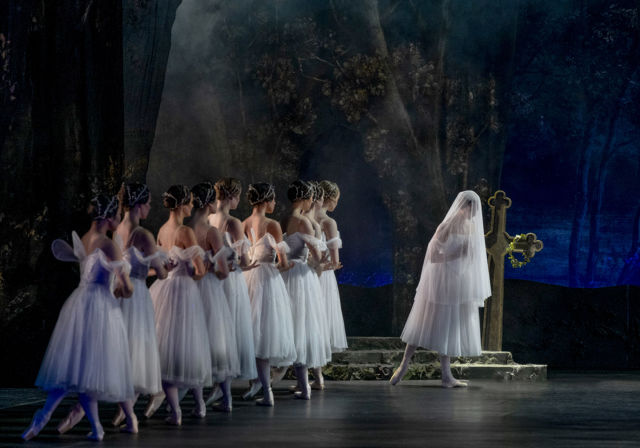 Ballettdansere i hvite tutuer står på mørk scene med skogsbakgrunn og blomsterkors, én danser har slør over ansiktet.