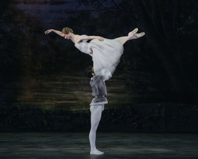 Ballettdanser løfter en annen danser horisontalt over hodet på mørk scene med skogsbakgrunn.
