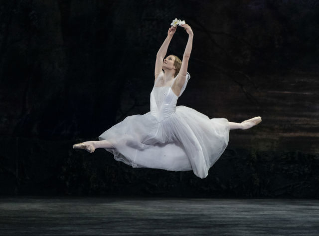 Ballettdanser i hvit tutu utfører et hopp med blomster i hendene på mørk scene.