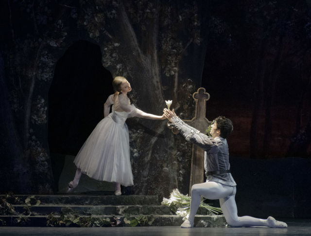 To ballettdansere på en mørk scene. Én står på en trapp i hvitt kostyme og tar imot blomster fra en knelende danser i grått. Bakgrunnen viser trær og et gravkors.
