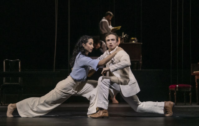 To dansere i dyp utfall mot hverandre på mørklagt scene, med en person som arbeider ved et skrivebord i bakgrunnen