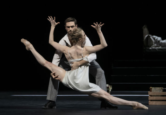 danser strekkes ut i en dyp, kontrollert posisjon mens partneren støtter bevegelsen på mørklagt scene