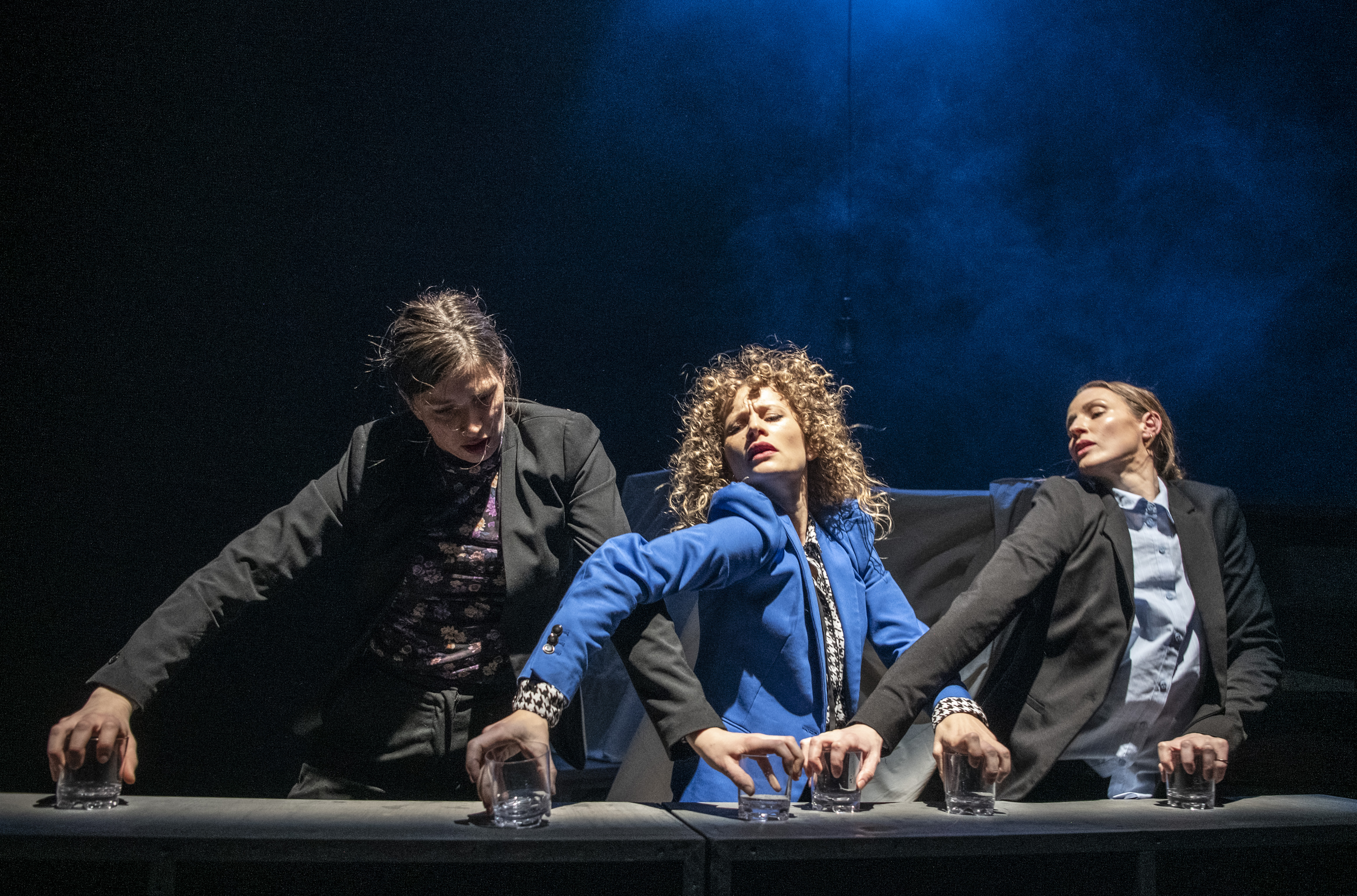 Tre personer på en teaterscene lener seg frem over et bord med glass, under dramatisk blått lys og røyk.