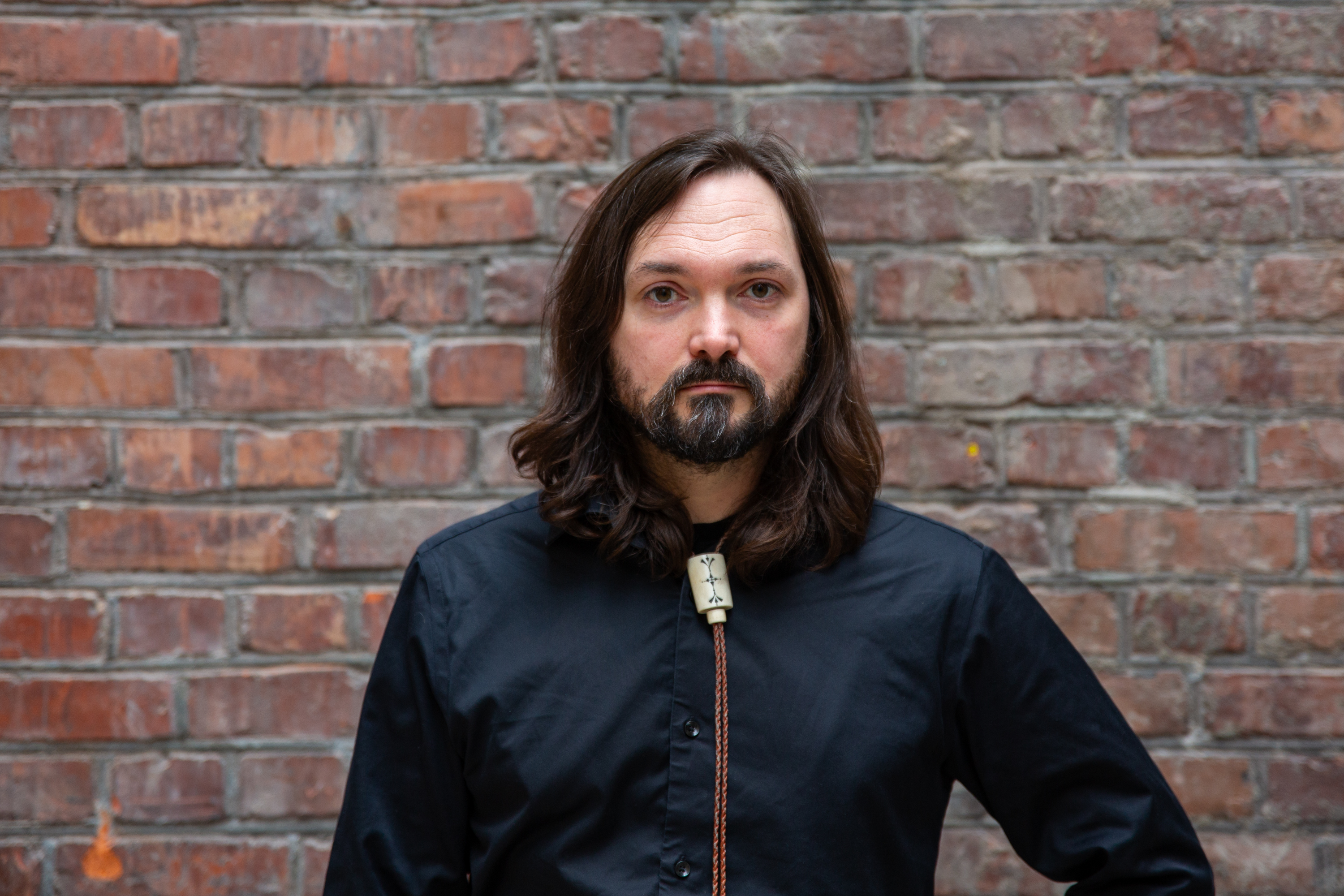 portrett av en mann med langt hår og skjegg foran en mursteinsvegg