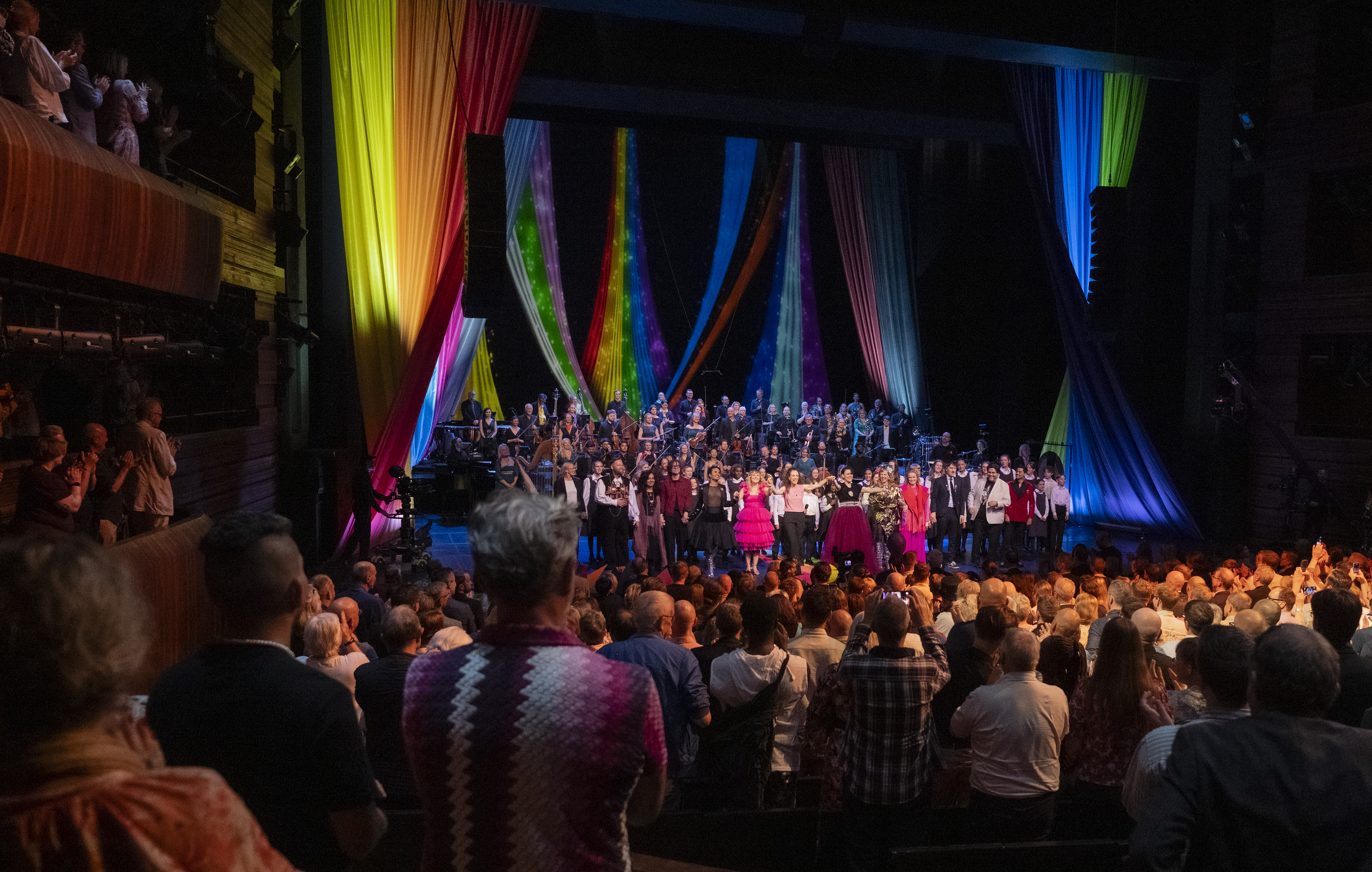 Stående publikum foran en mengde utøvere på scene, under pride-dekorasjoner