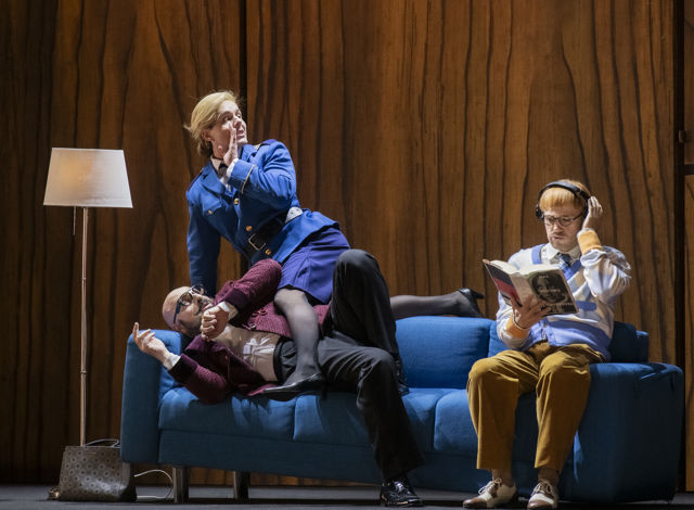 Kvinne sitter delvis oppå mann på sofa, en annen mann sitter ved siden av og leser