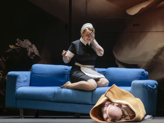 Kvinne utkledd som tjenestepike sitter på sofa og ler, mens mann er rullet inn i teppe på gulvet