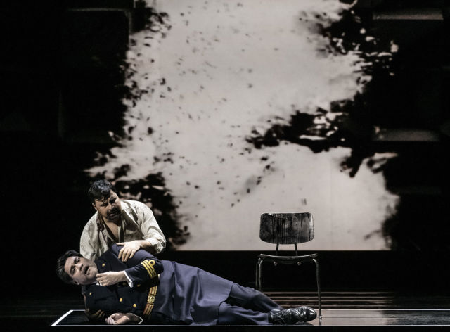 Scenebilde av mann i militæruniform som ligger såret på bakken, annen mann på kne bak ham