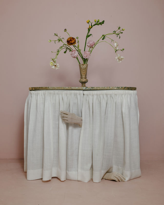 Et bord med en vase med blomster. To hender kommer til syne gjennom et gardin under bordet.