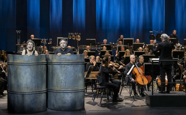 En kvinne og en mann i store tønner foran orkester på en scene
