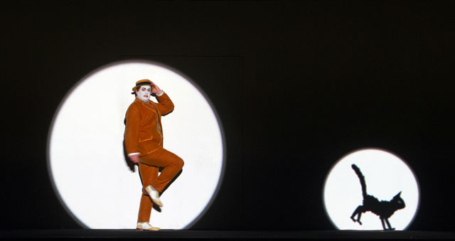 Buster Keaton-lignende mann i rød dress i sirkulært spotlys på scene, med katt foran seg.