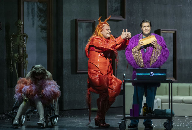 Tre rollefigurer på scenen ved et bord, en er i rødt djevelkostyme